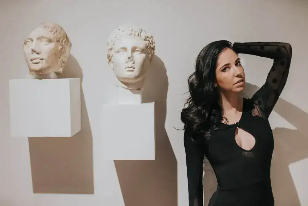 A woman posing beside two classical marble bust sculptures.