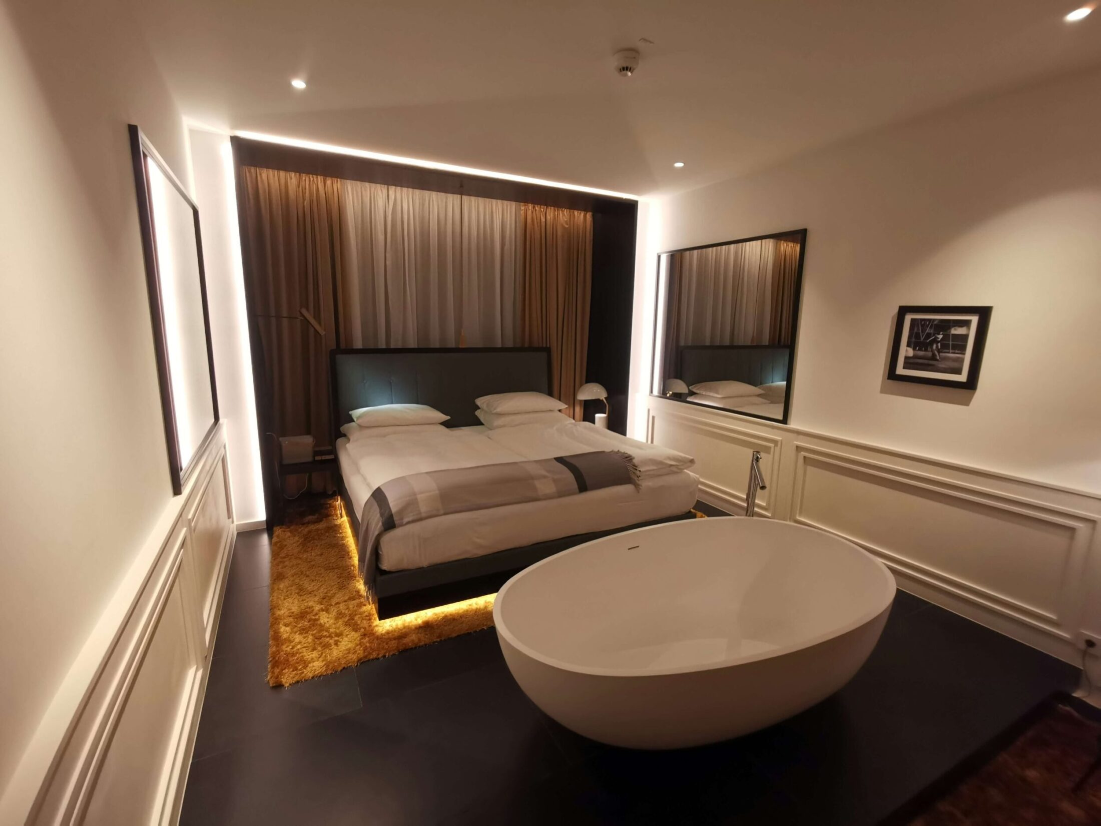 Modern luxury hotel suite with double bed and oval bathtub.